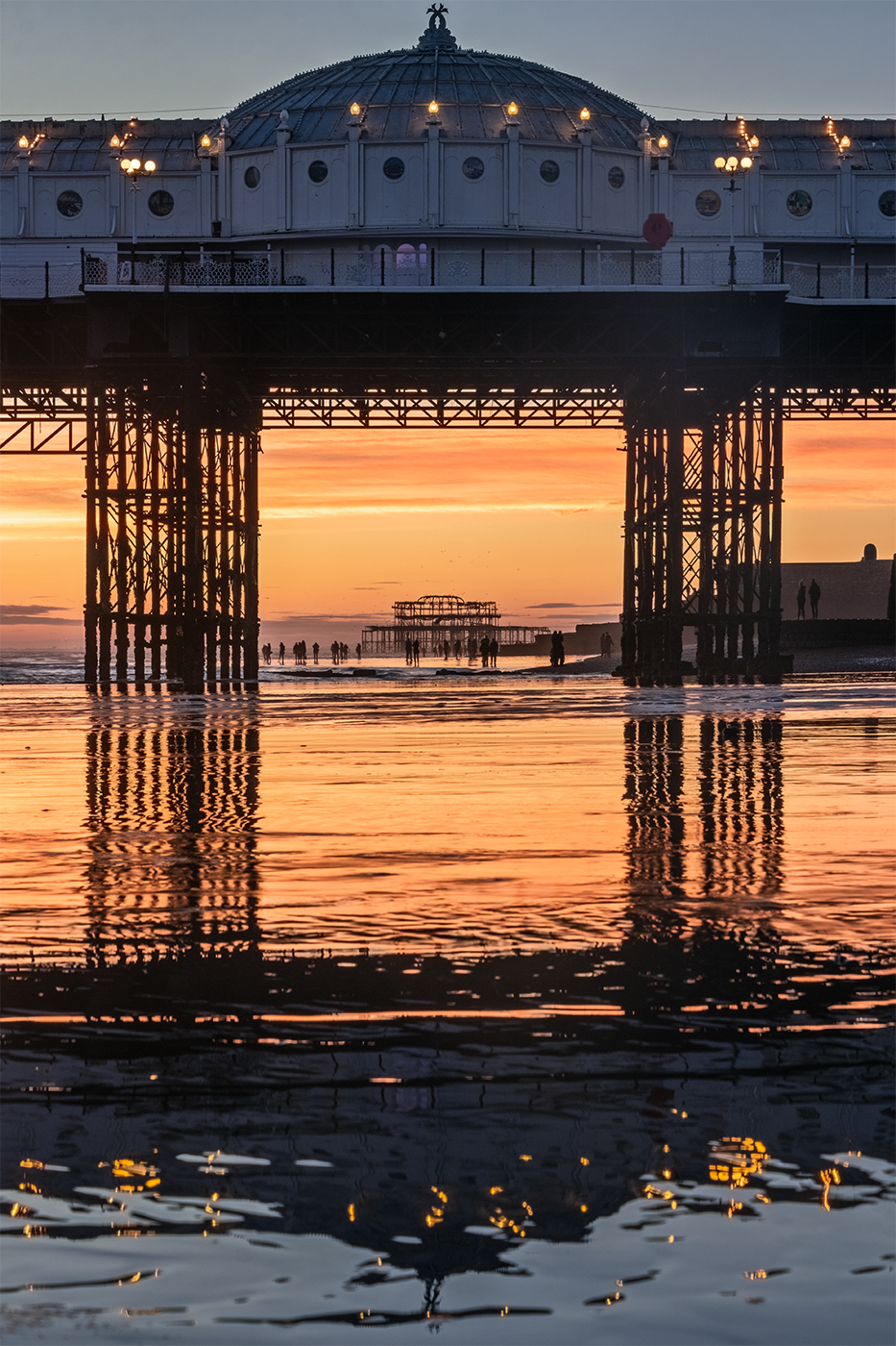 Brighton pier is reflected in the wet sand as the sun sets along the shoreline at low tide.