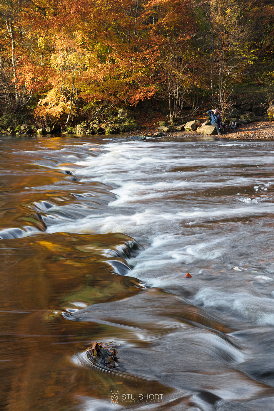 A person relaxes momentarily on a rock, gazing into the low sun against an autumnal backdrop of vibrant trees as a wide river cascades in the foreground.