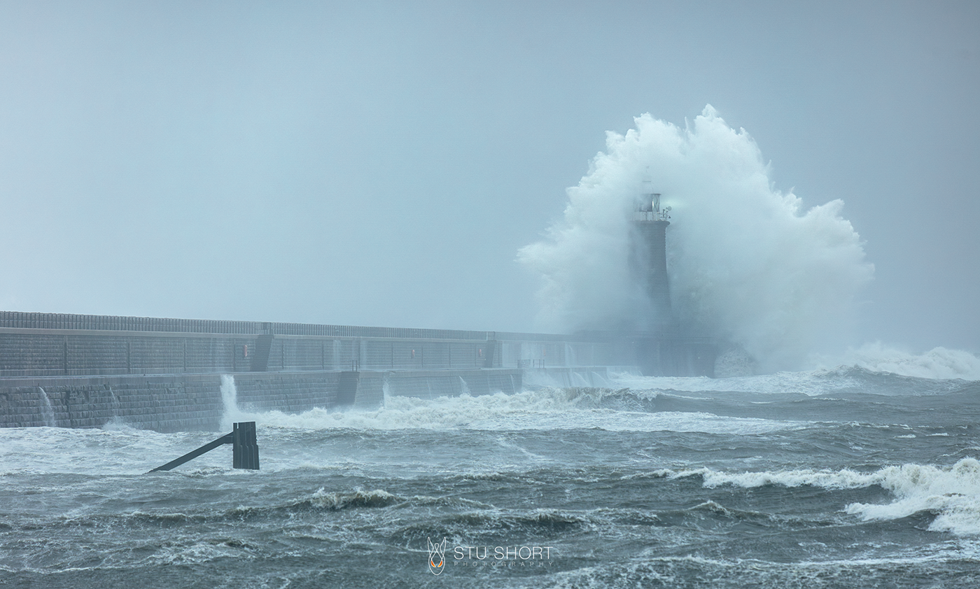 Tynemouth lighthouse is enveloped by tumultuous waves, symbolizing safety and guidance in the face of nature's fury.