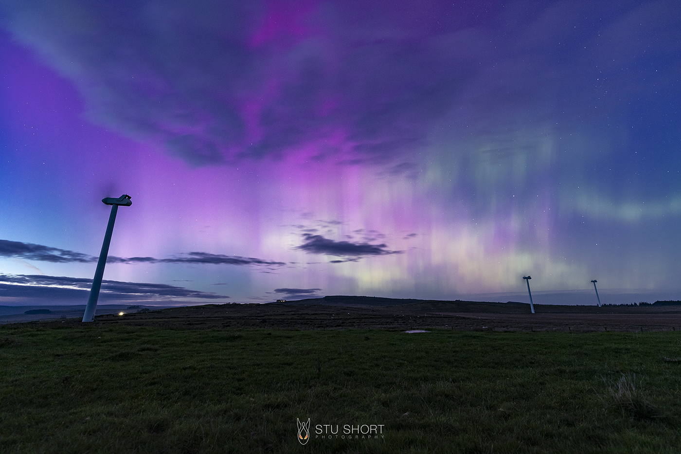 A stunning view of the aurora borealis illuminating a windfarm sat in the Northumberland countryside with vibrant colors against a night sky.