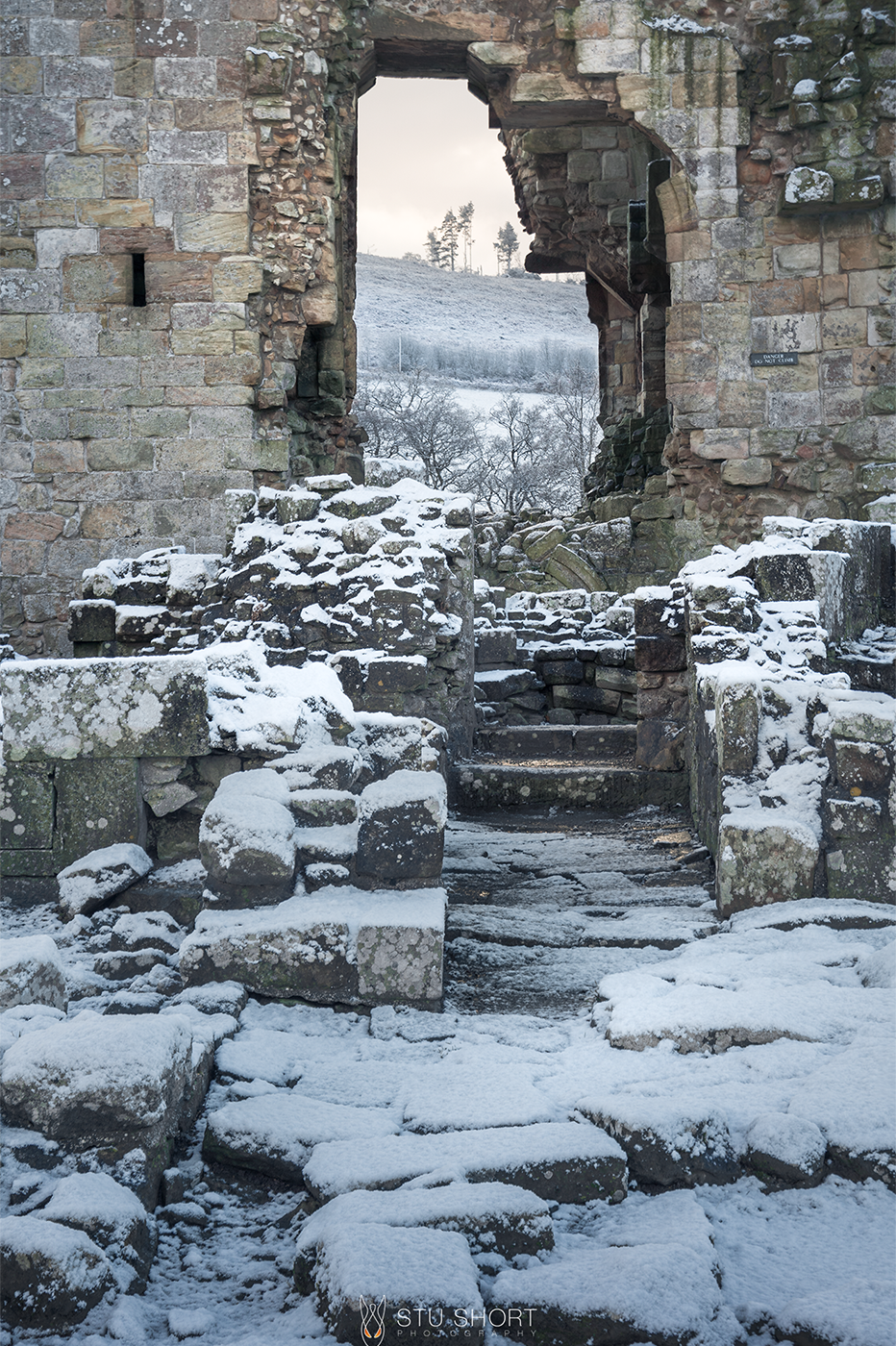 A dusting of snow covers the ruins that lead to the open doorwar of Edlingham castle and the snow covered hills outside.