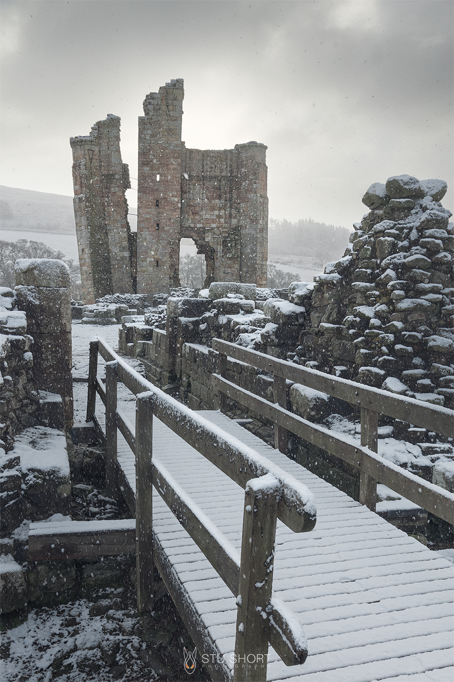 A snow covered footbridge leads into the grounds of a stunning ruined castle which looms tall in the distance, creating a fairytale scene.