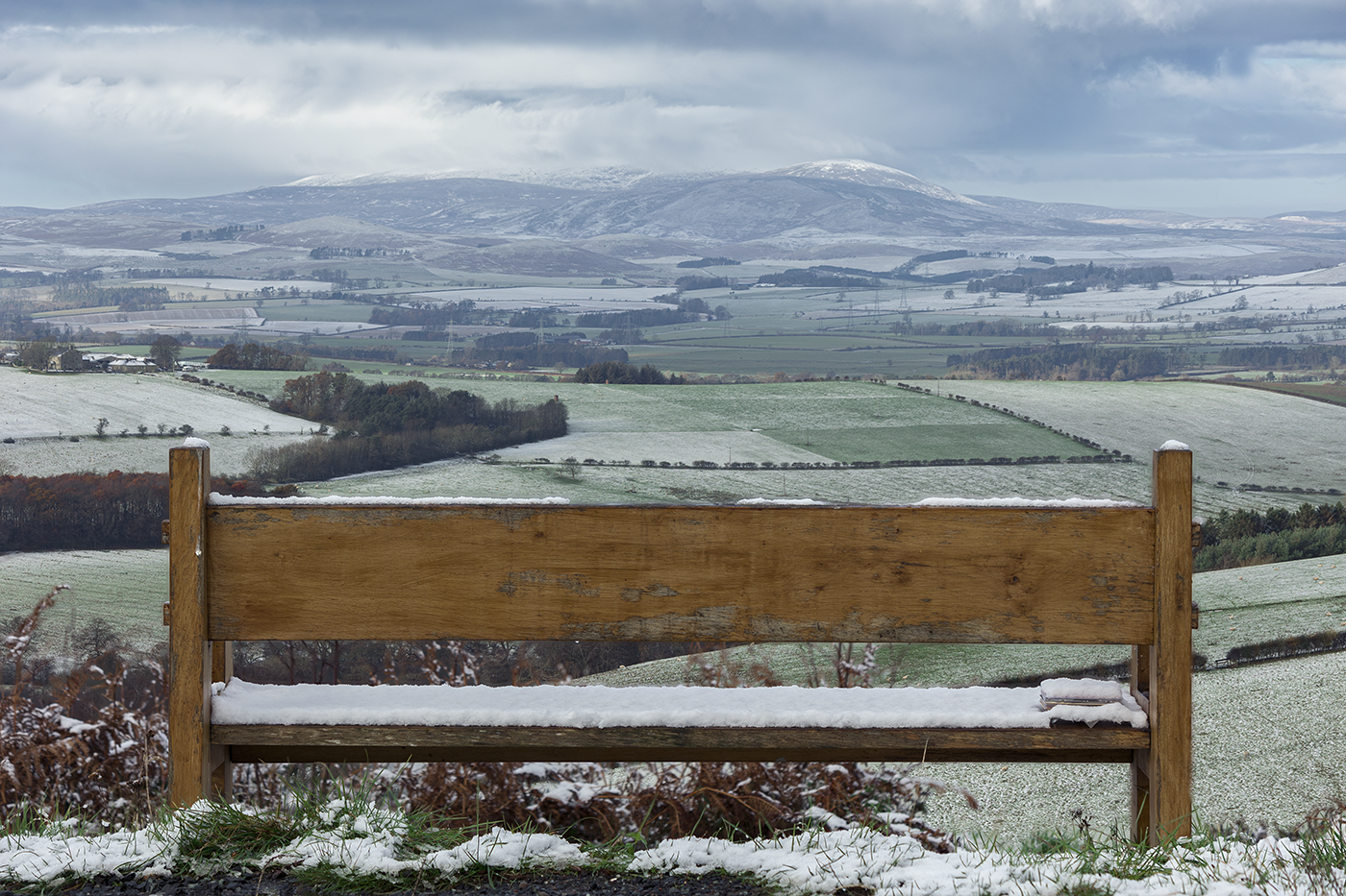 Winter landscape photography featuring a snow covered bench overlooks a serene landscape of snow covered rolling hills.