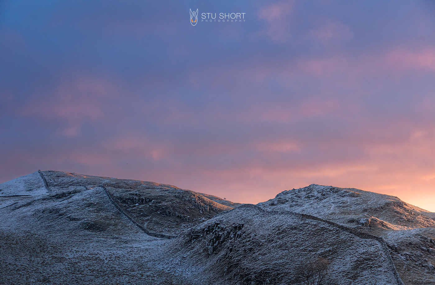 Winter landscape photography featuring the meandering path of Hadrian's Wall crests the tops of a series of hills that reaches up into a glorious sunrise.