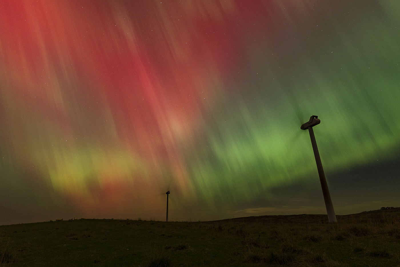 Two wind turbines are silhouetted against a vibrant, fiery northern lights display.