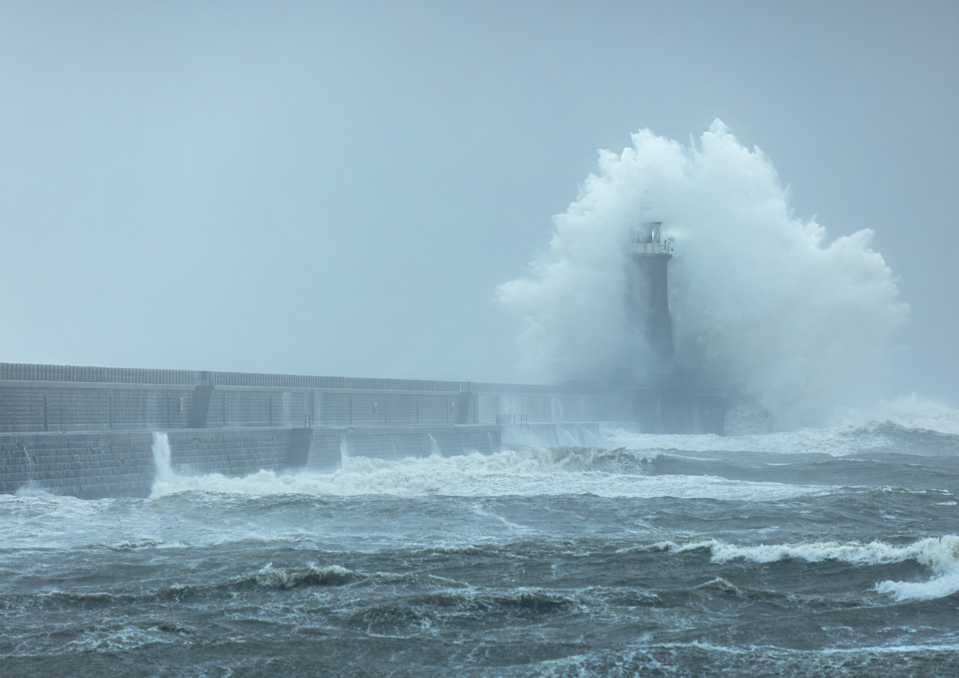 Huge waves crashing over a lighthouse and pier during a storm.