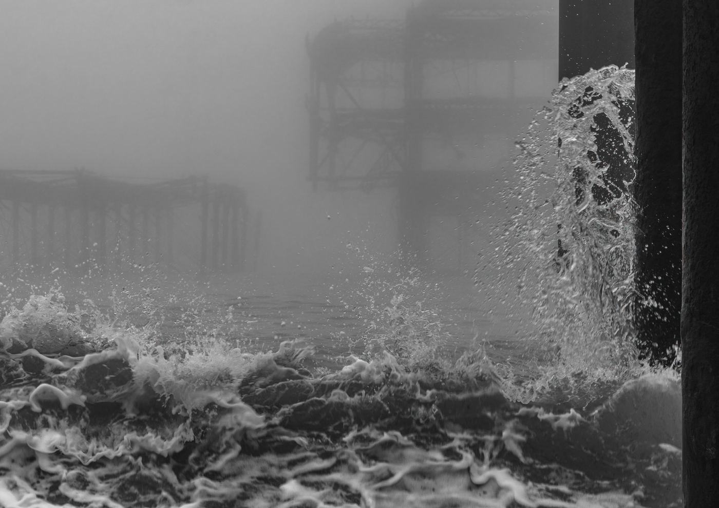 Black and white image of waves crashing against a pier with a foggy background.