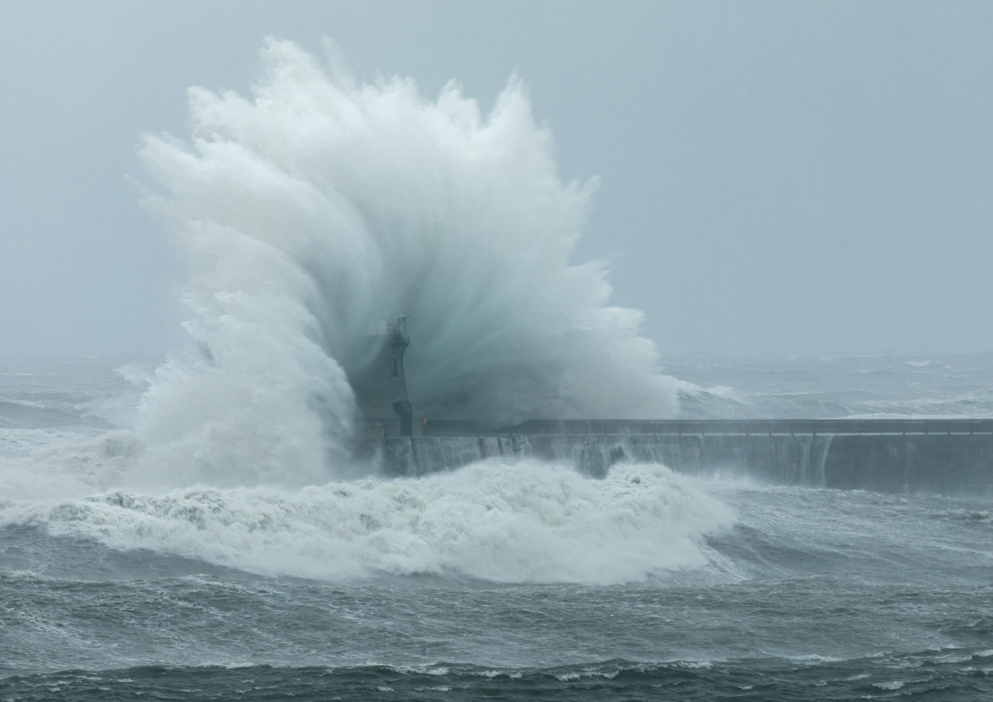 Powerful wave engulfing a lighthouse with spray and foam exploding upwards.