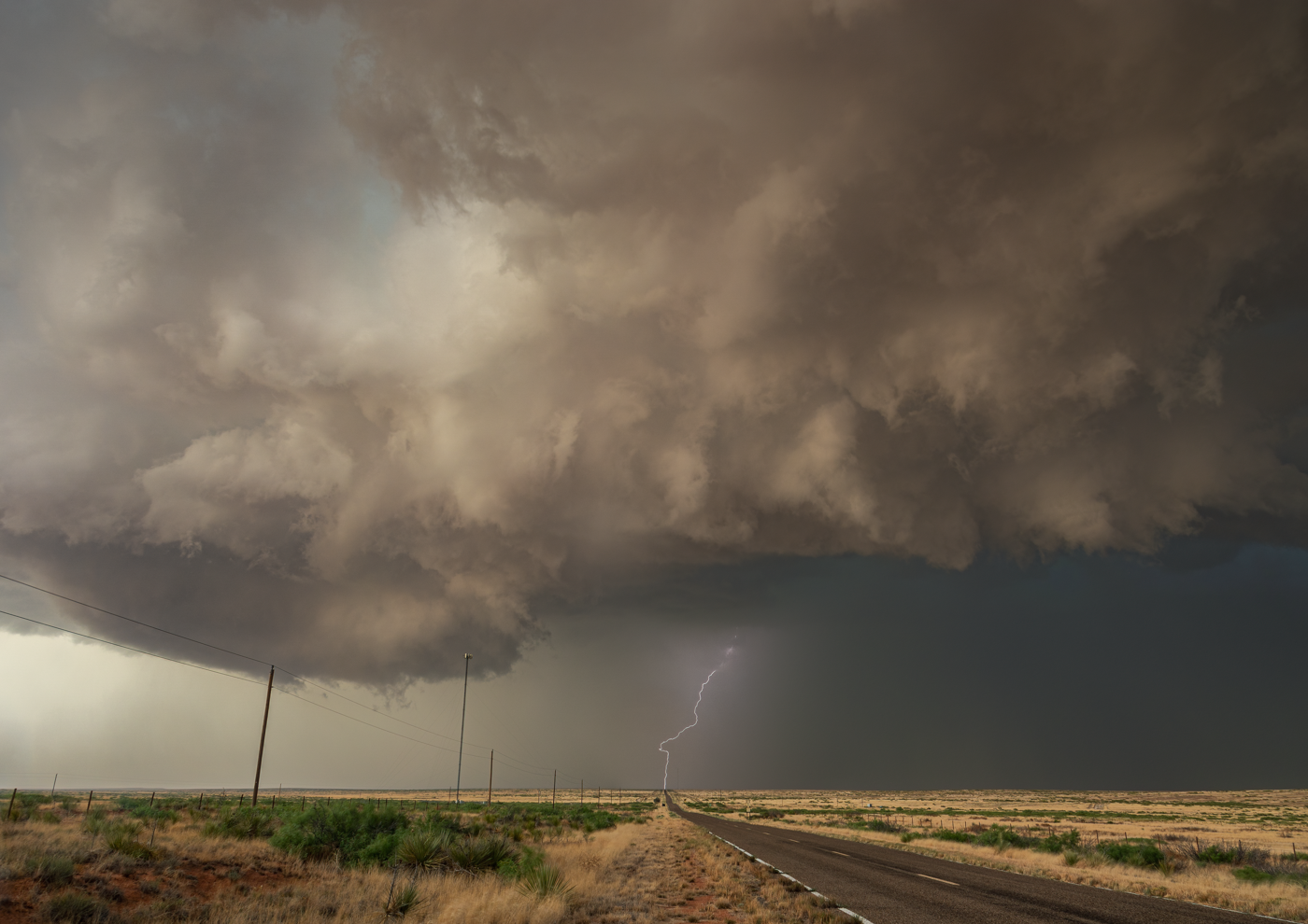 A large, ominous storm cloud dominates the sky over a long, empty highway, with a visible lightning strike and dark rain falling in the distance.