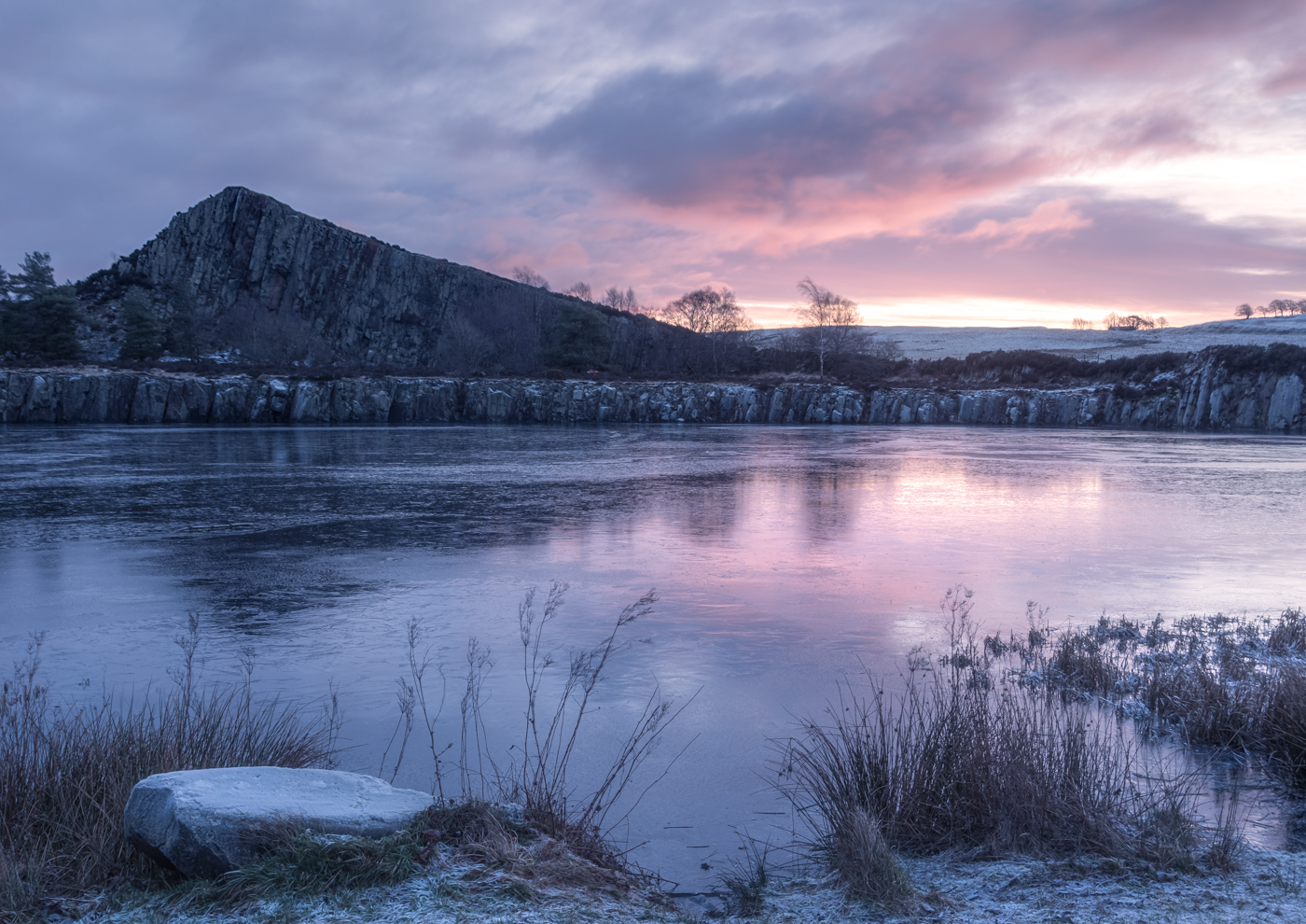 A serene winter landscape with a frozen pool reflecting a pastel-colored sunset sky, framed by a dark, craggy rock face on one side and snow-covered ground in the foreground.