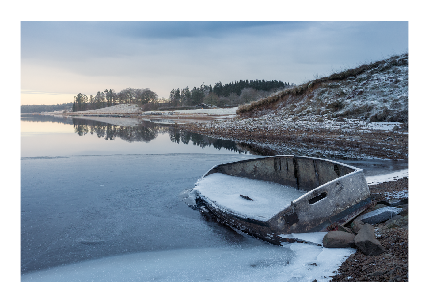 Abandoned boat frozen in the ice on a still lake with snow-covered trees in the distance