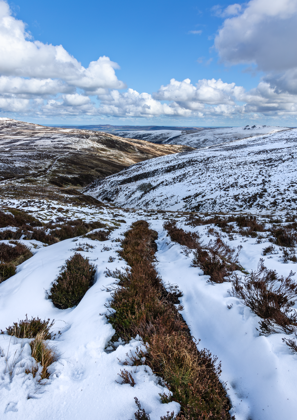 A snowy moorland landscape with rolling hills, patches of snow, brown heather, and a bright blue sky with fluffy white clouds.